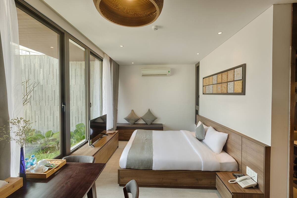A modern hotel room with a large bed, a wooden desk, a dining table, and a view through the sliding glass door.