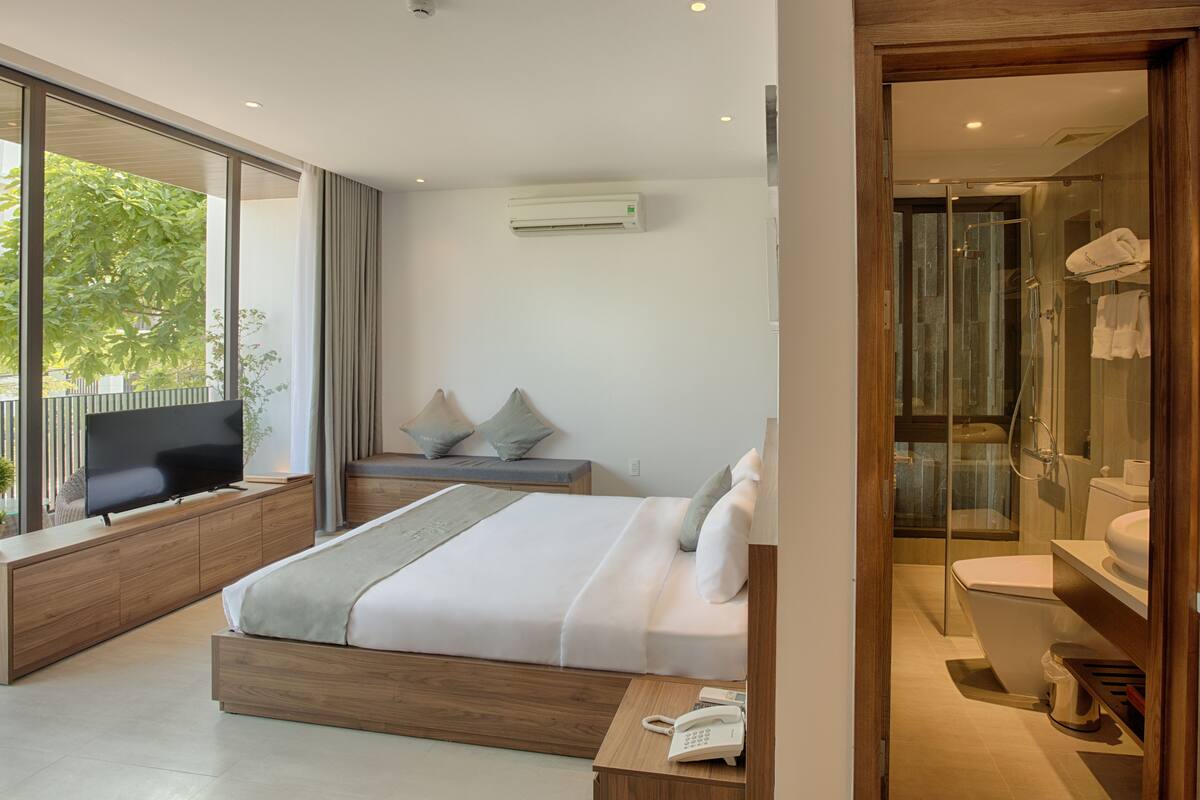 A modern hotel room with a large bed, a TV, a bedside table, and a glass-enclosed shower area.