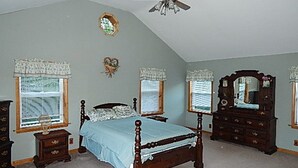 A bedroom with a large bed, two nightstands, a dresser, a ceiling fan, and two windows with shutters.