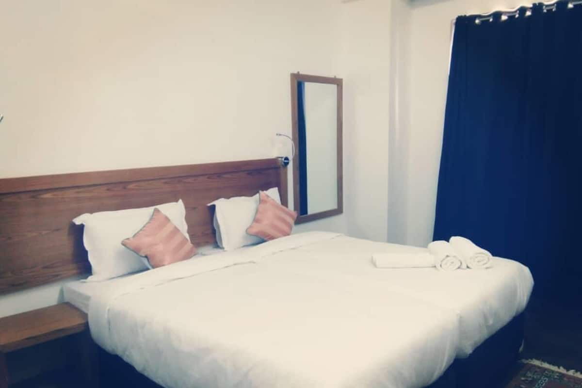 A neatly made bed with white bedding and pink pillows, a wooden headboard, a bedside table, and a mirror on the wall.