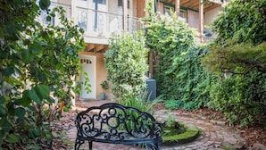 Un banc situé dans un espace jardin, avec un chemin en briques, entouré de verdure et d’un bâtiment en arrière-plan.