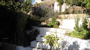 Un escalier en pierre mène à un bâtiment aux murs blancs, et un jardin agrémenté de diverses plantes se trouve à proximité.
