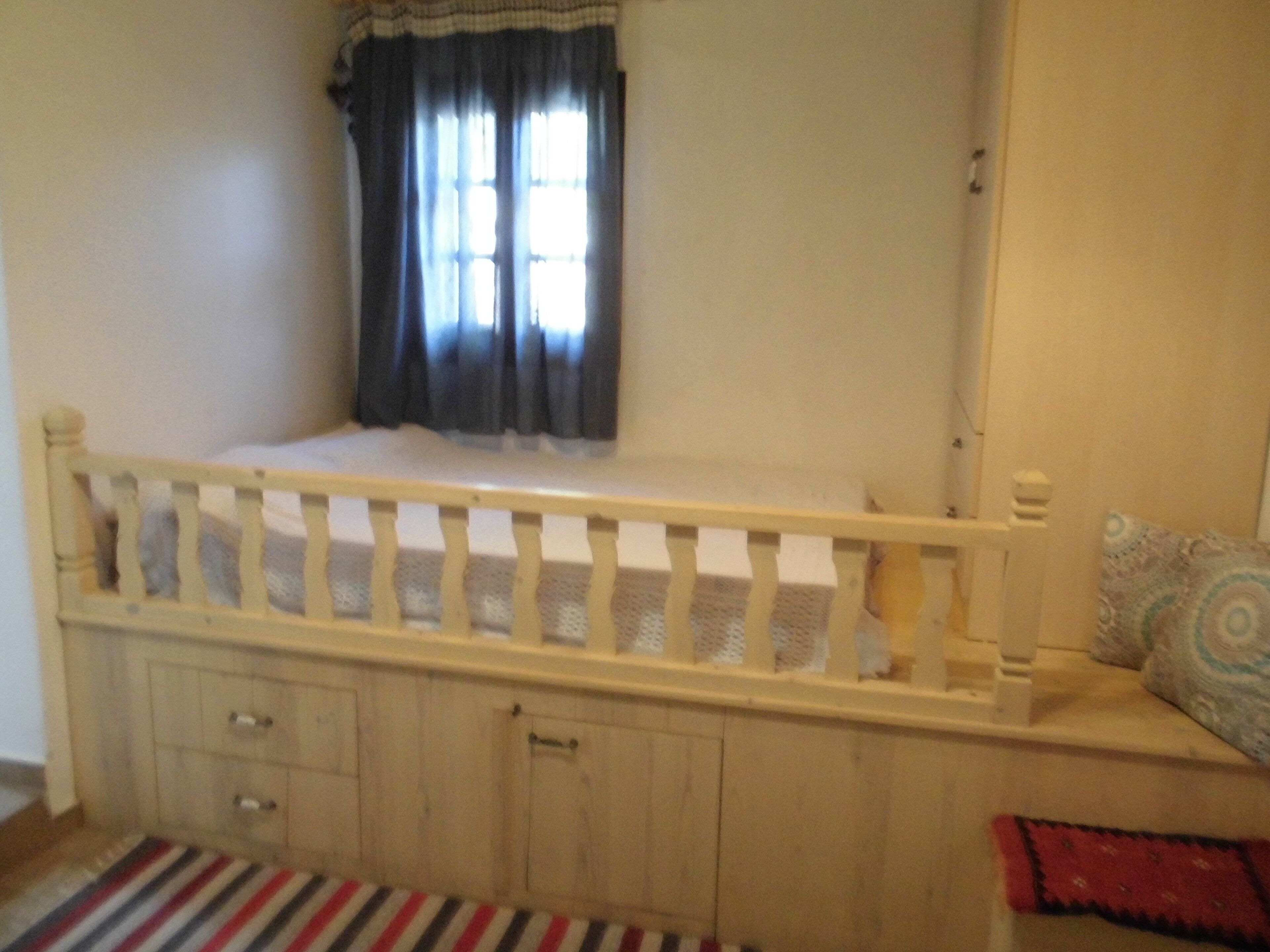 Quarto com estrutura de cama de madeira, janela com cortinas, mesa de cabeceira e tapete listrado.