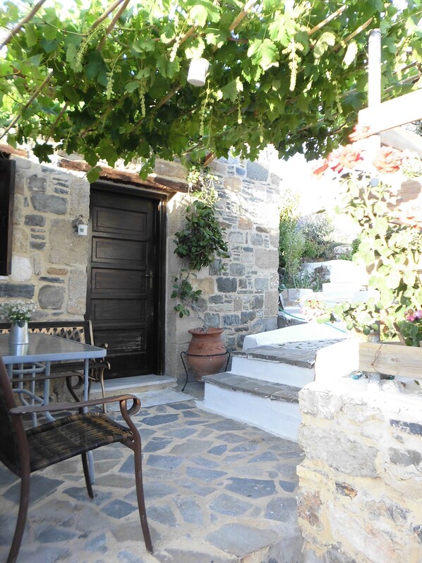 A stone building with a dark wooden door, a potted plant, a table with chairs, and a stone floor.