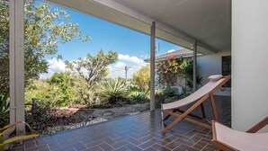 Eine überdachte Terrasse mit gefliestem Boden, zwei Liegestühlen und einem Garten mit verschiedenen Pflanzen und einem kleinen Wasserspiel.