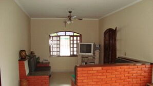 A living room with a TV, a brick accent wall, a ceiling fan, and a window with bars. - House with pool, barbecue, air conditioning. (Caraguatatuba)
