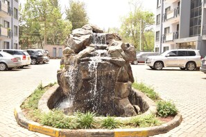 Una fuente circular con agua en cascada rodeada de una zona pavimentada, con coches aparcados y edificios al fondo.