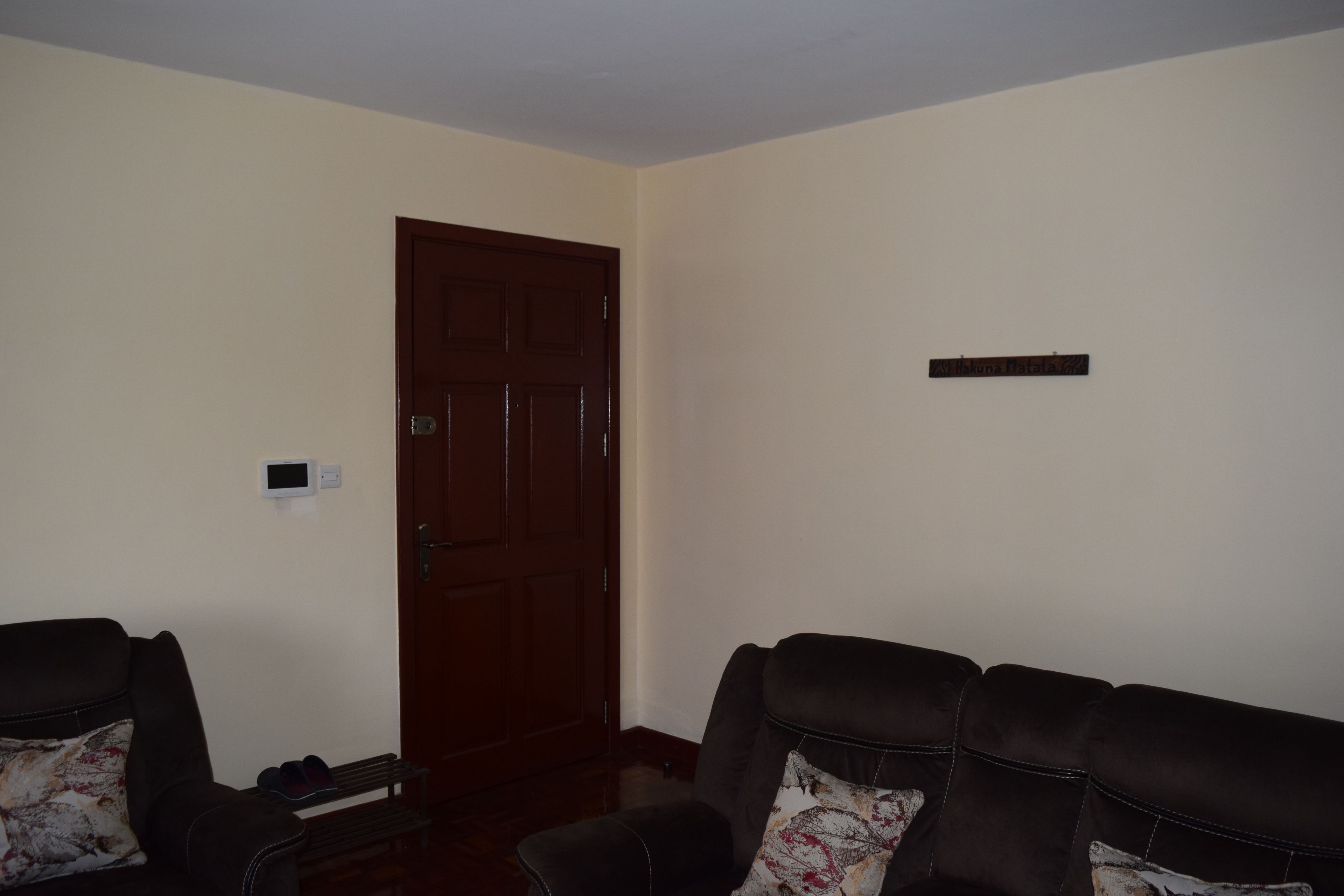 A room with a brown door, two black leather sofas with patterned cushions, and a small shelf with shoes.