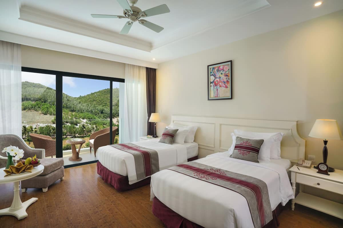 A hotel room with two beds, a ceiling fan, a view of greenery, a painting, and a table with flowers.