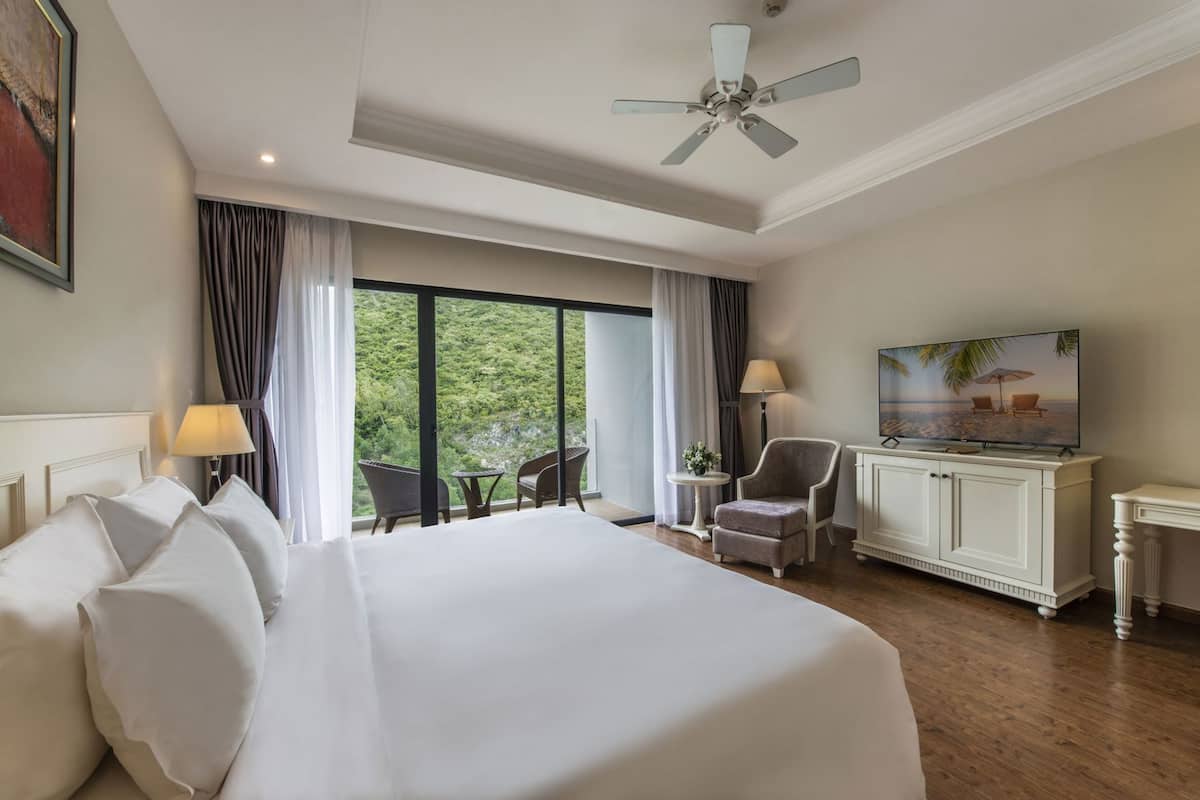 A hotel room with a large bed, a television, a ceiling fan, a view of greenery, and a balcony with chairs.