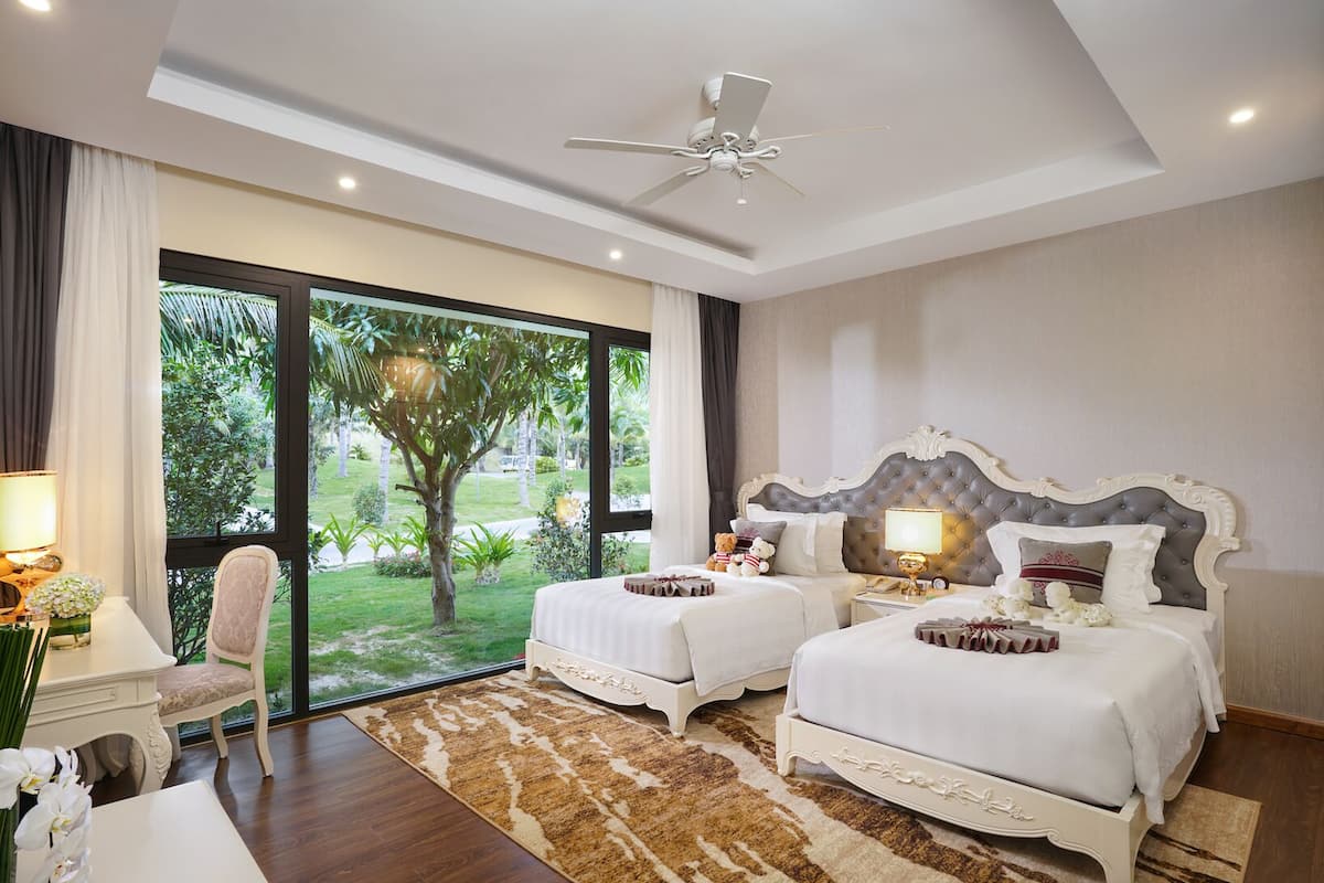 A hotel room with two beds, a ceiling fan, a large window with a view of greenery, and a small table with a lamp.