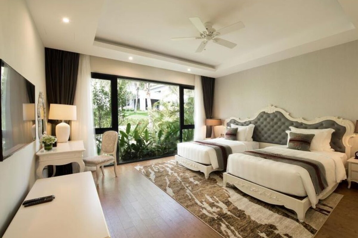 A hotel room with a bed, bedside tables, a desk, a chair, a ceiling fan, and a view of greenery outside.