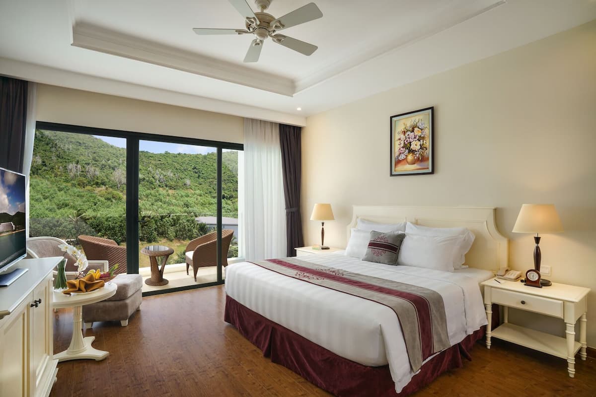 A hotel room with a bed, two bedside tables, a TV, a desk, a chair, and a balcony with a view of greenery.