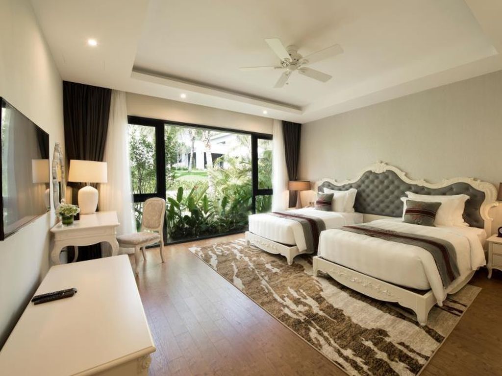 A hotel room with a bed, bedside tables, a desk, a chair, a ceiling fan, and a view of greenery outside.