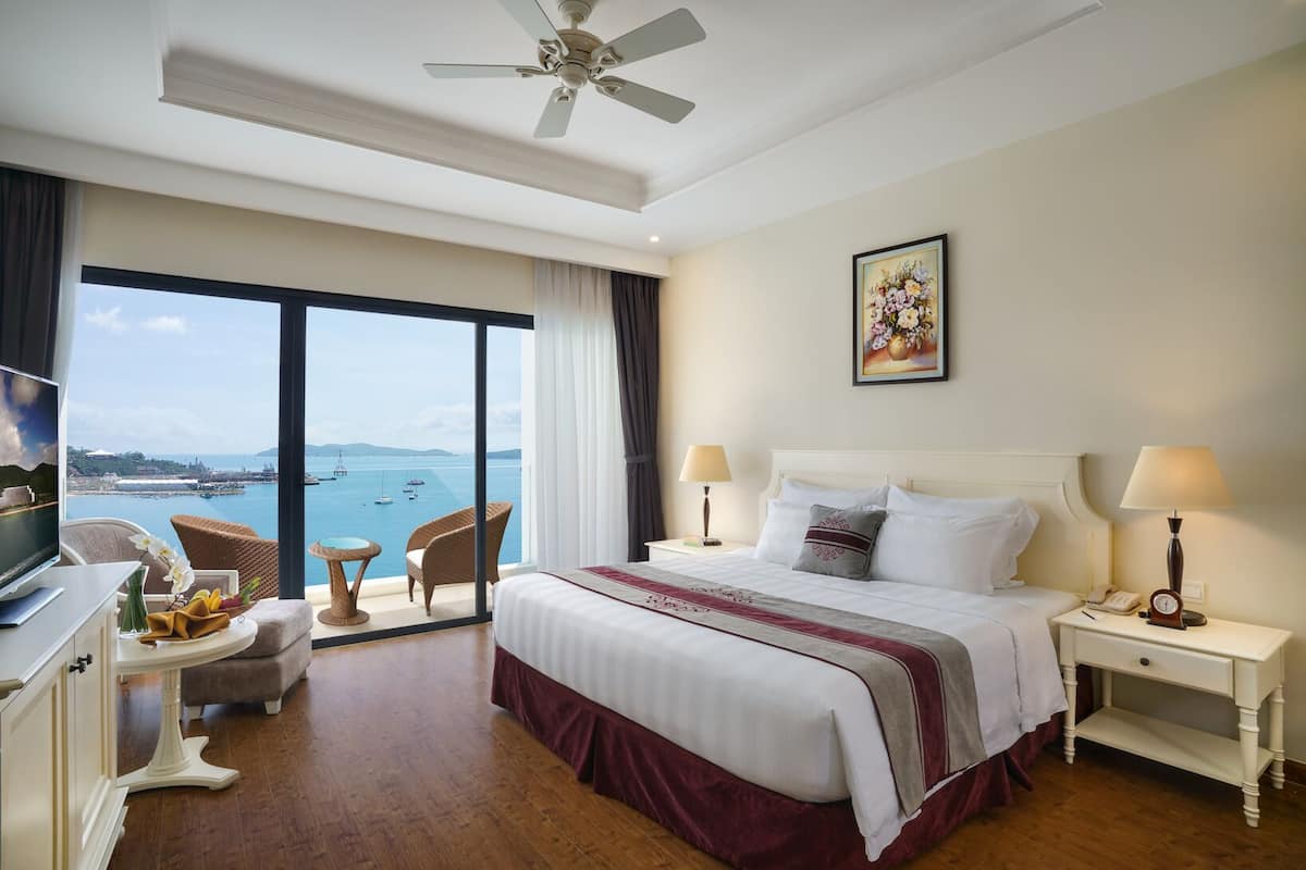 A hotel room with a bed, two bedside tables, a TV, a chair, a small table, and a large window with a view of the sea.