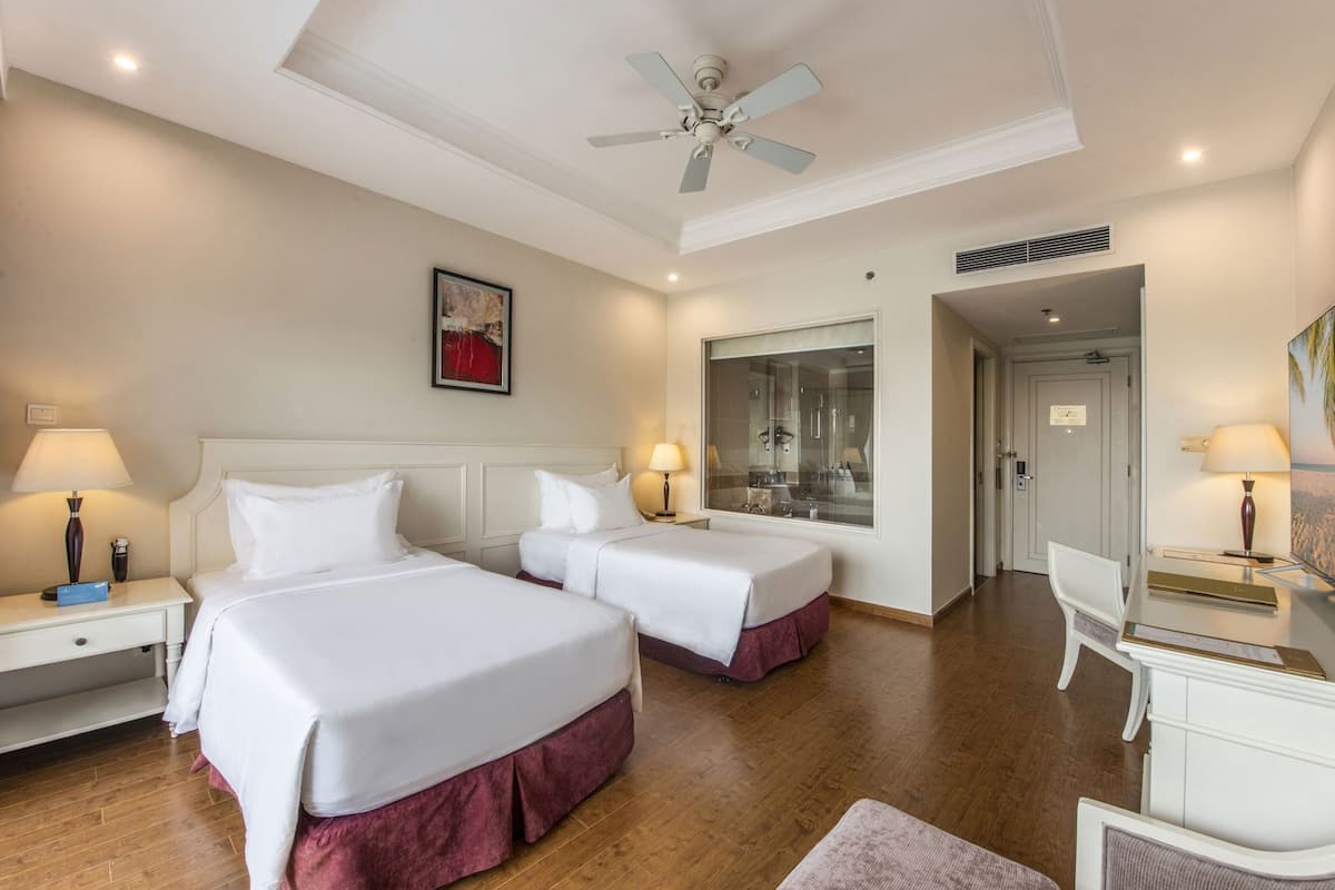 A hotel room with two beds, a desk, a chair, a ceiling fan, and a large window.