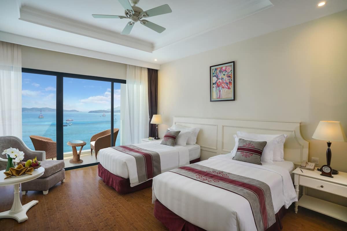 A hotel room with two beds, a ceiling fan, a large window with a view of the sea, and a painting on the wall.