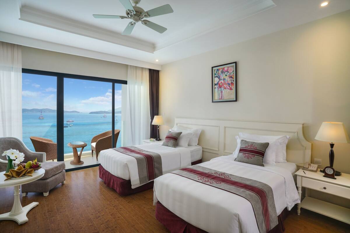 A hotel room with two beds, a ceiling fan, a large window with a view of the sea, and a painting on the wall.