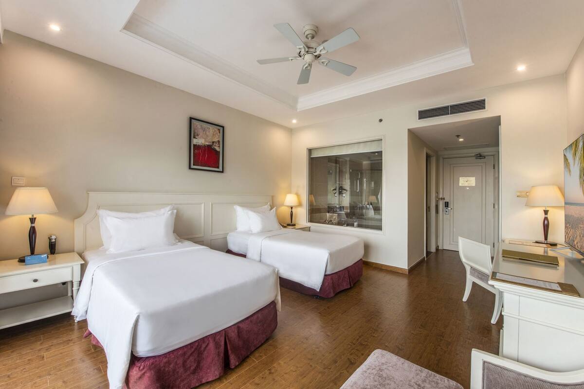 A hotel room with two beds, a desk, a chair, a ceiling fan, and a large window.