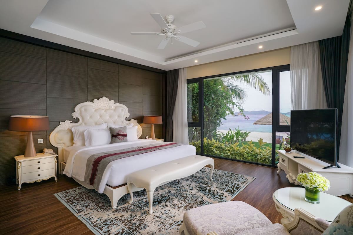 A modern bedroom with a large bed, a ceiling fan, a TV, and a view of the beach.