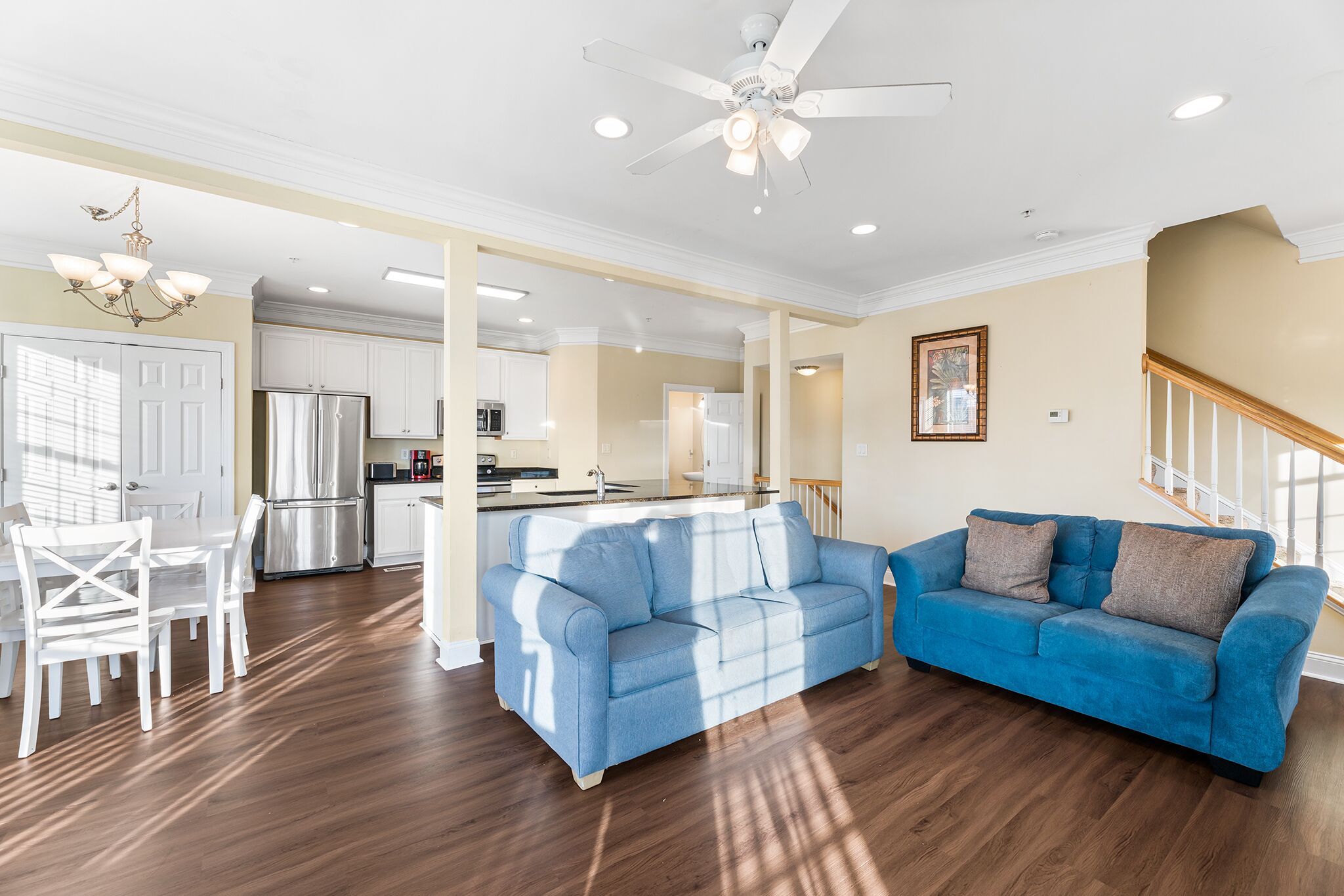 A spacious living room with a blue sofa, white dining table, and chairs. A kitchen with white cabinets and stainless steel appliances is visible in the background.