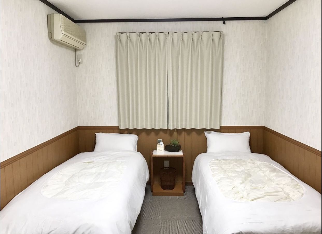 Habitación de hotel con dos camas individuales, cabecero de madera, una mesita de noche pequeña con lámpara y una ventana con cortinas.