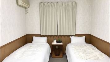 Habitación de hotel con dos camas individuales, cabecero de madera, una mesita de noche pequeña con lámpara y una ventana con cortinas.
