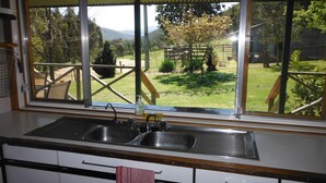 A kitchen with a large window, two sinks, and a view of a grassy area with trees and a wooden structure. - Riverbend Homestead Horse and Farmstay (Freeburgh)