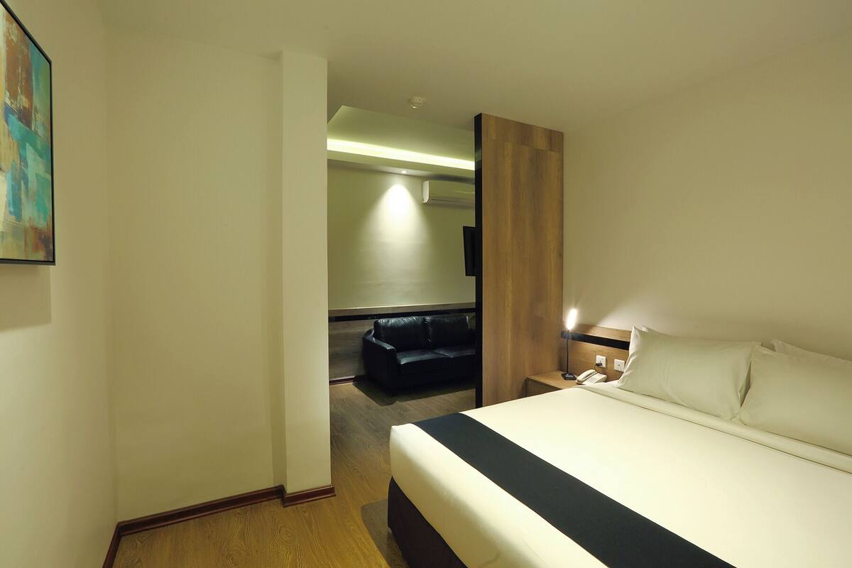 A hotel room with a bed, a nightstand, a lamp, a wall-mounted TV, and a door leading to a sitting area with a sofa.