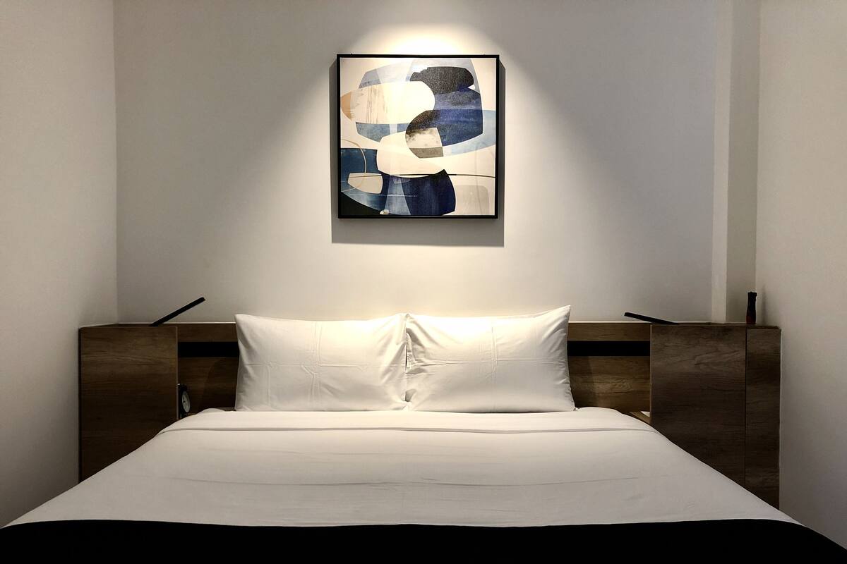 A neatly made bed with white bedding and a black accent at the foot. A modern abstract painting hangs above the bed.