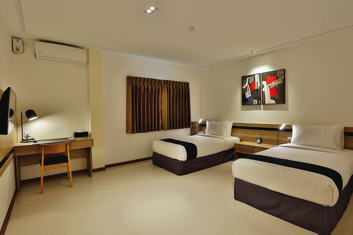 A hotel room with two beds, a desk, a chair, an air conditioner, a clock, and artwork on the wall.