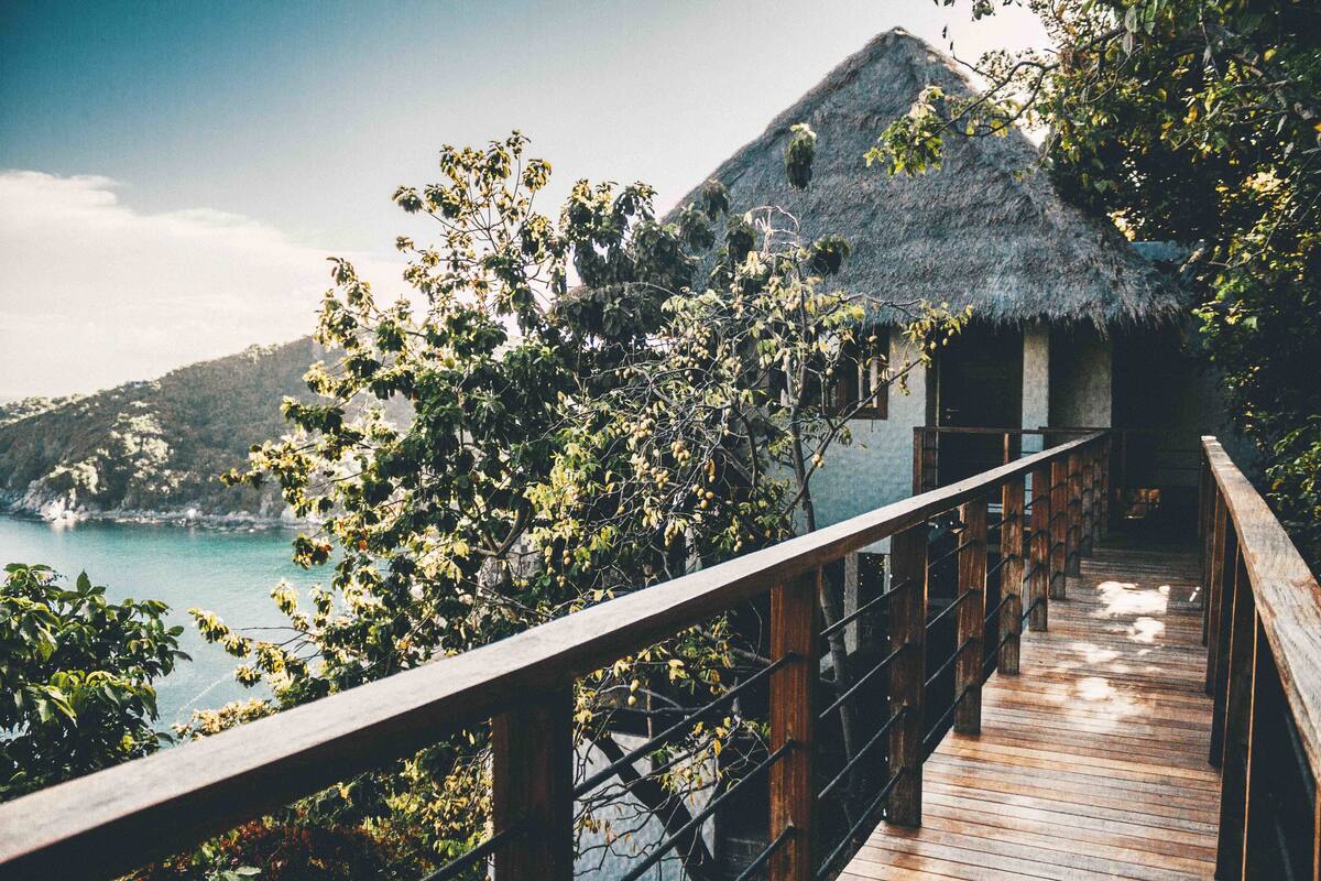 Une terrasse en bois mène à un bâtiment dont le toit est recouvert de chaume, et qui offre une vue sur un paysage côtier.