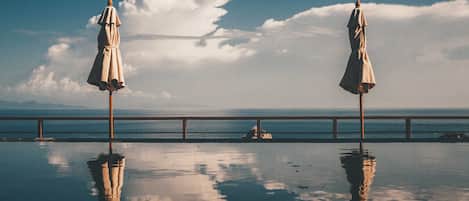 Two beach umbrellas on a reflective pool with a railing and a view of the sea and clouds.