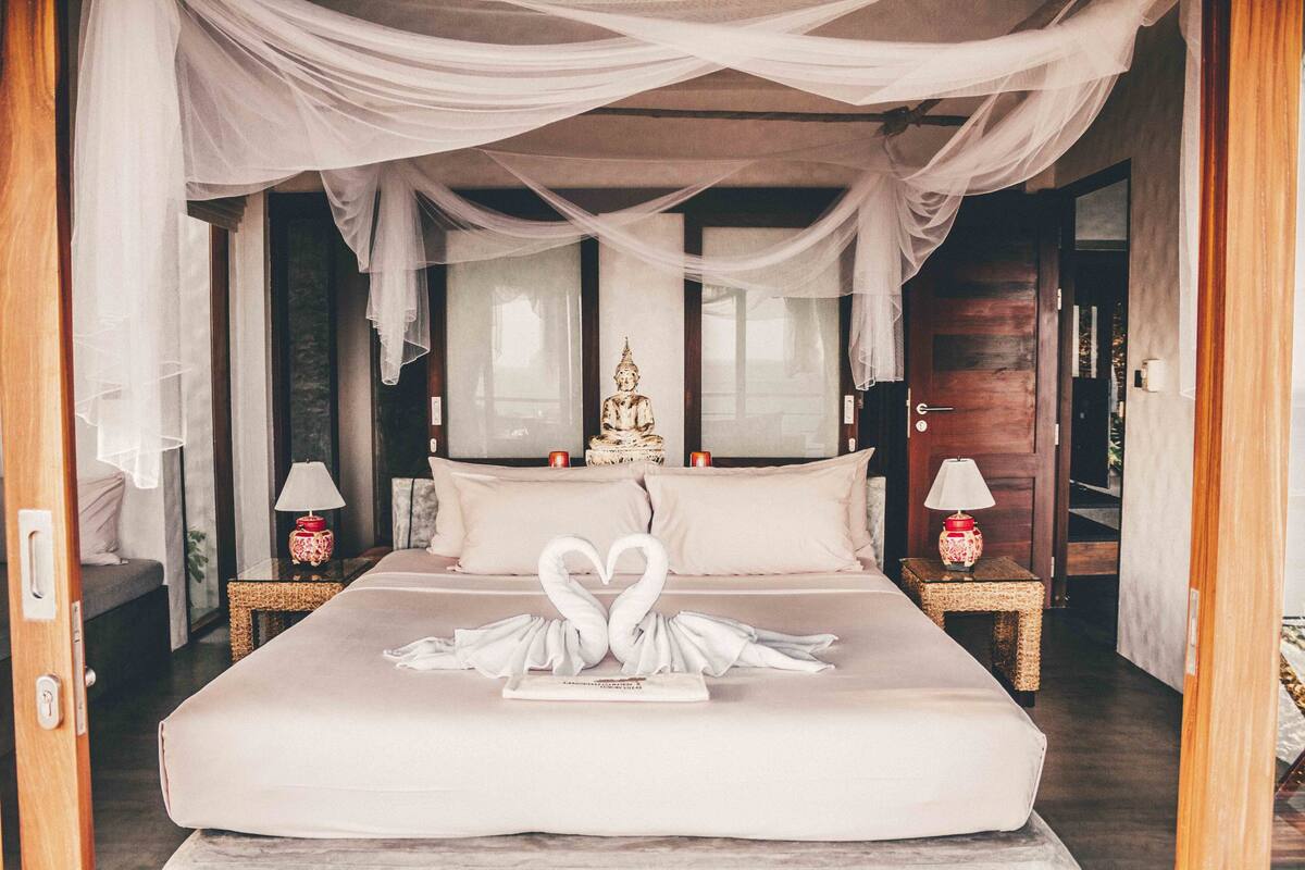 Une chambre à coucher avec un lit, deux tables de chevet, une statue de Bouddha et une décoration en forme de cœur faite avec des serviettes.