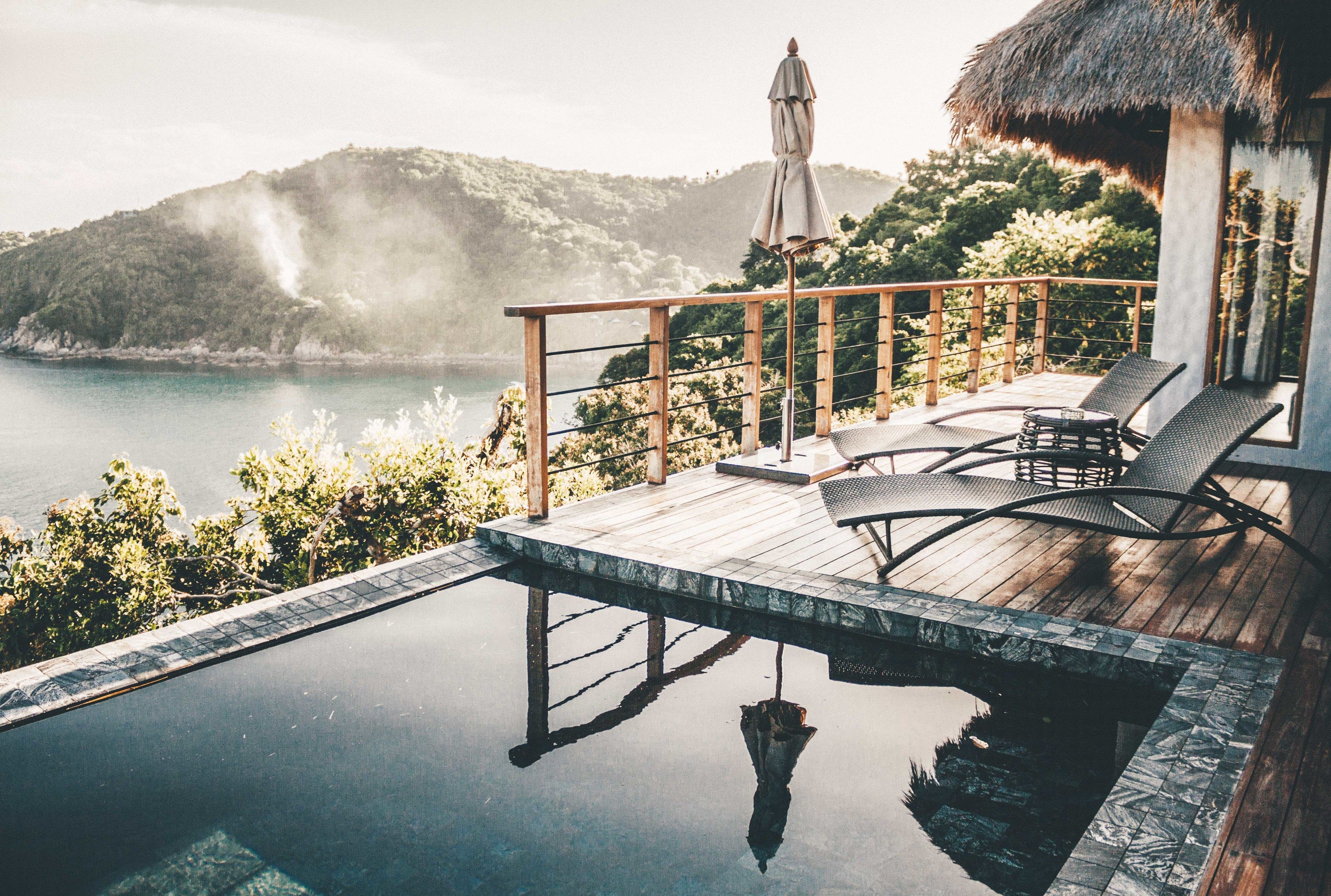An infinity pool with a wooden deck, lounge chairs, and a thatched-roof structure overlooking a body of water and hills.