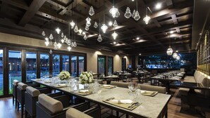 Restaurante com uma longa mesa de jantar posta para uma refeição, rodeado de cadeiras e lustres de cristal pendurados.