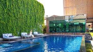 Piscina com espreguiçadeiras, um edifício de vidro e uma parede verde.