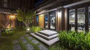 Fachada de madeira com entrada por porta de vidro, pequena área externa com mesa e cadeiras, e gramado bem cuidado com pedras para pisar.