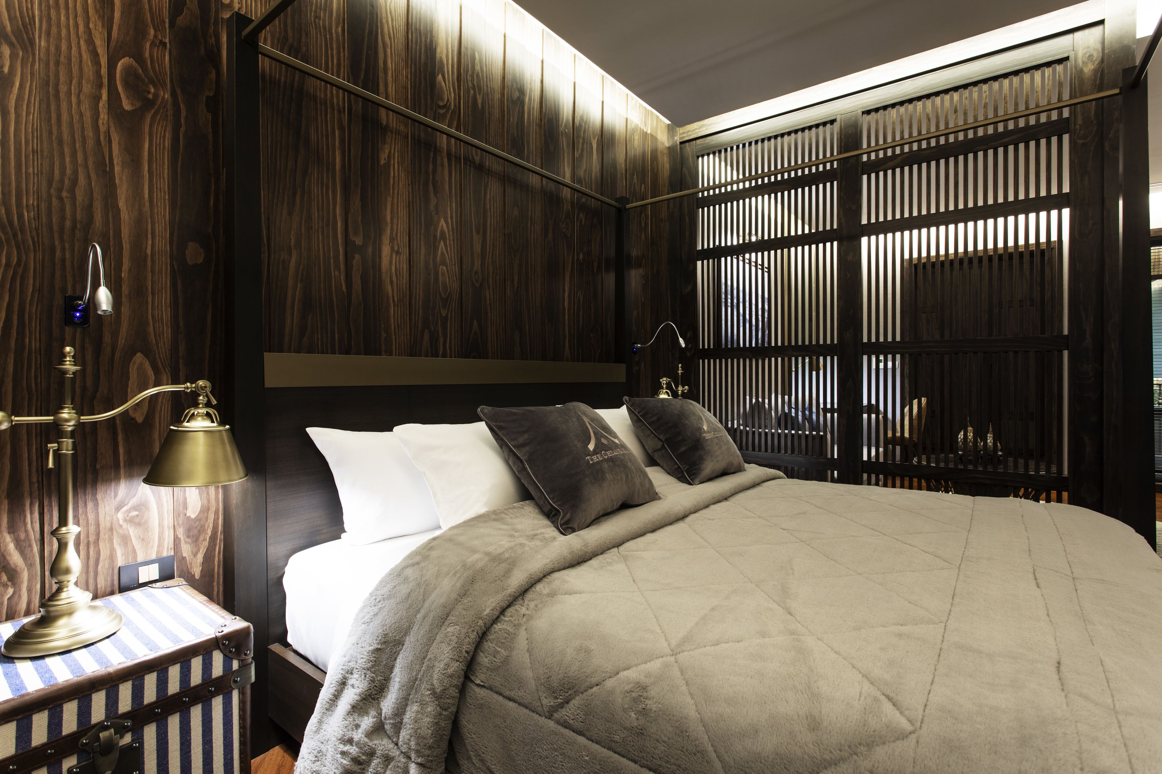 A bedroom with a wooden headboard, a bed with a quilted cover, two pillows, a nightstand with a lamp, and a window with blinds.