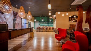 Eine geräumige Lobby mit einem hölzernen Empfangsbereich, roten Stühlen, hängenden Pendelleuchten und einer Bar im Hintergrund.
