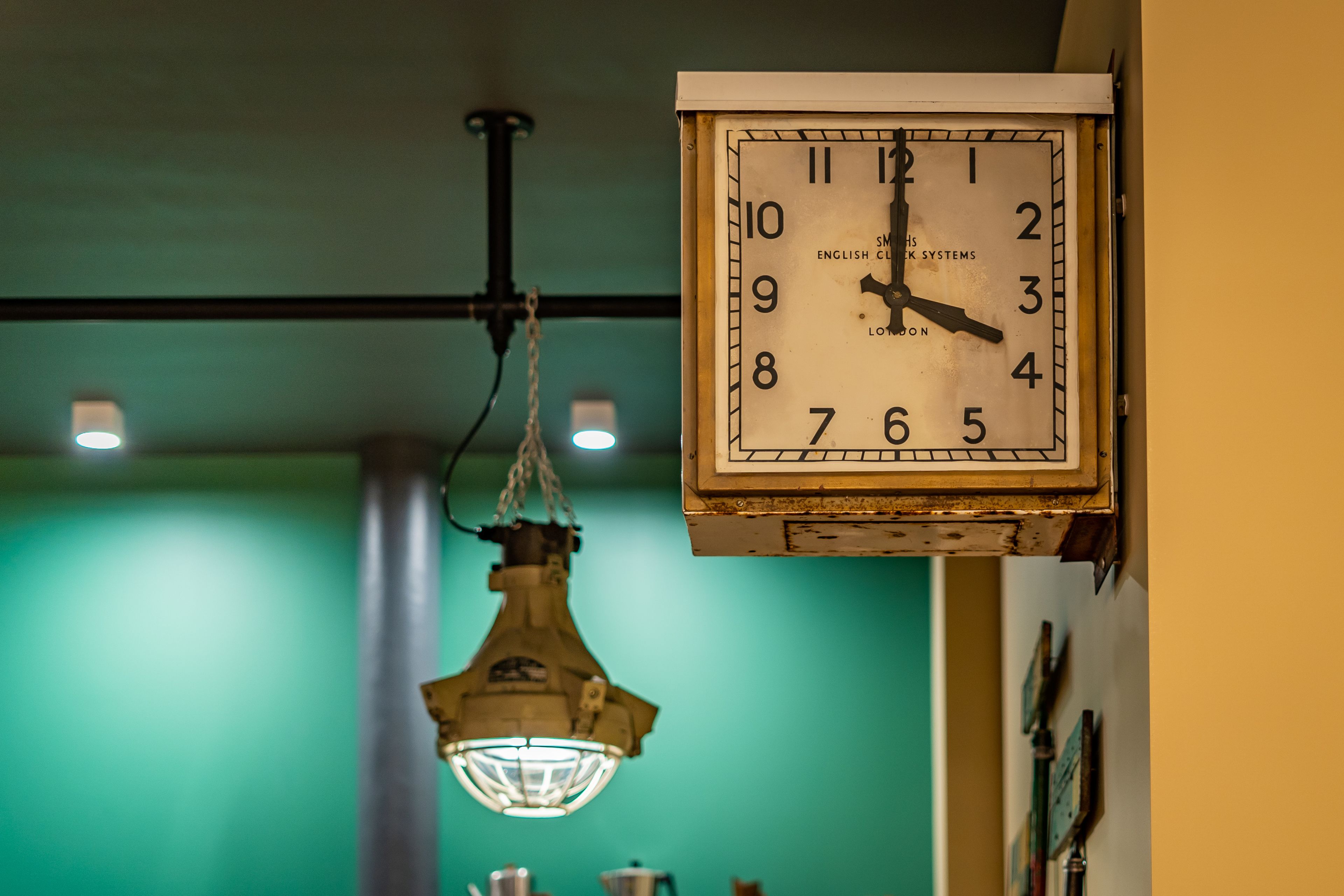 Un reloj de pared con diseño clásico, una lámpara colgante de estilo industrial y una pared verde al fondo.