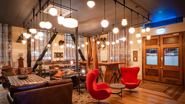Eine großzügige Lobby mit Holzboden, Ledersofas, einem roten Sessel und mehreren Pendelleuchten.