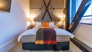 Ein modernes Hotelzimmer mit einem großen Bett, einem markanten, X-förmigen Kopfteil, zwei Nachttischlampen und Blick durch ein Fenster.