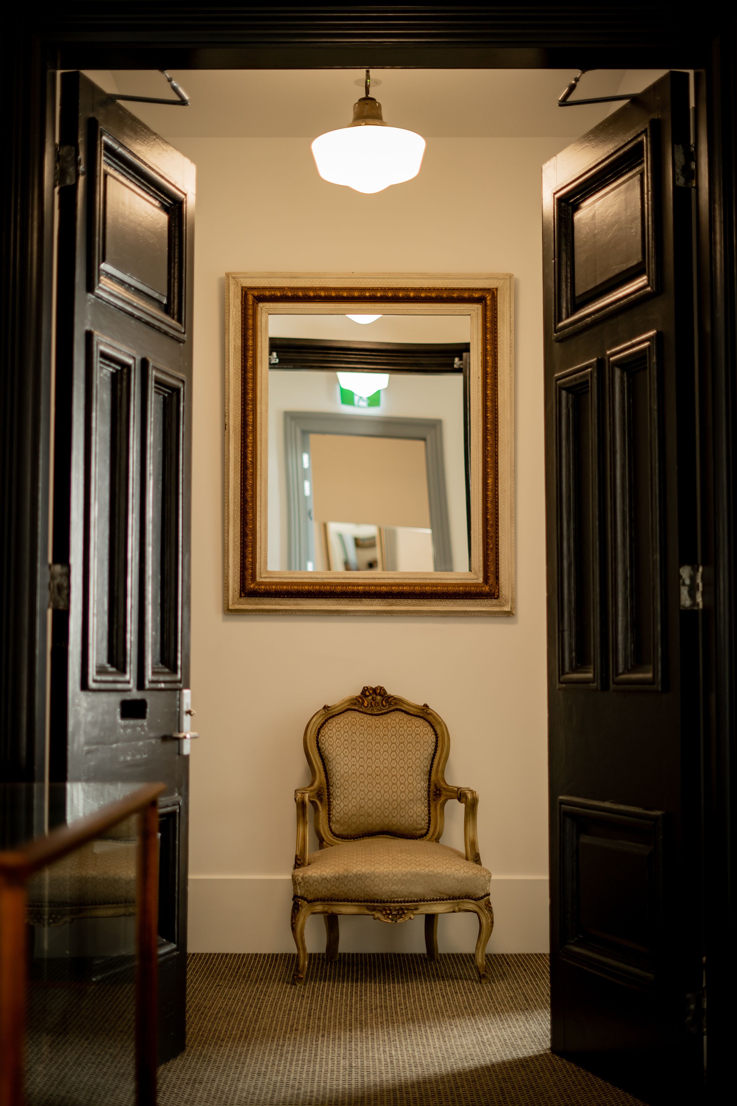 A hallway with a framed mirror, a chair, and dark wooden doors.