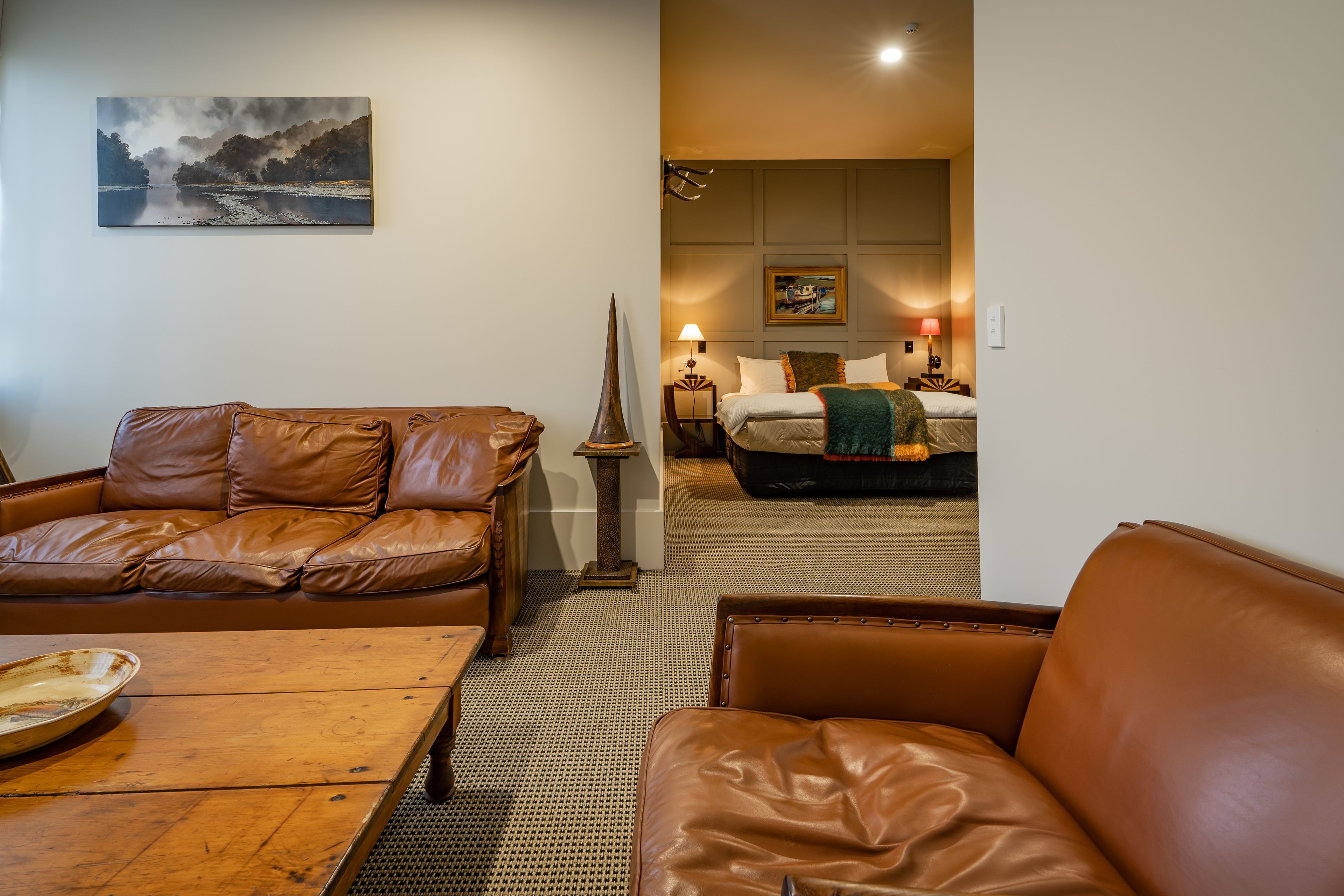 A hotel room with a brown leather sofa, a wooden coffee table, a cat tree, and a bed with a green throw blanket.