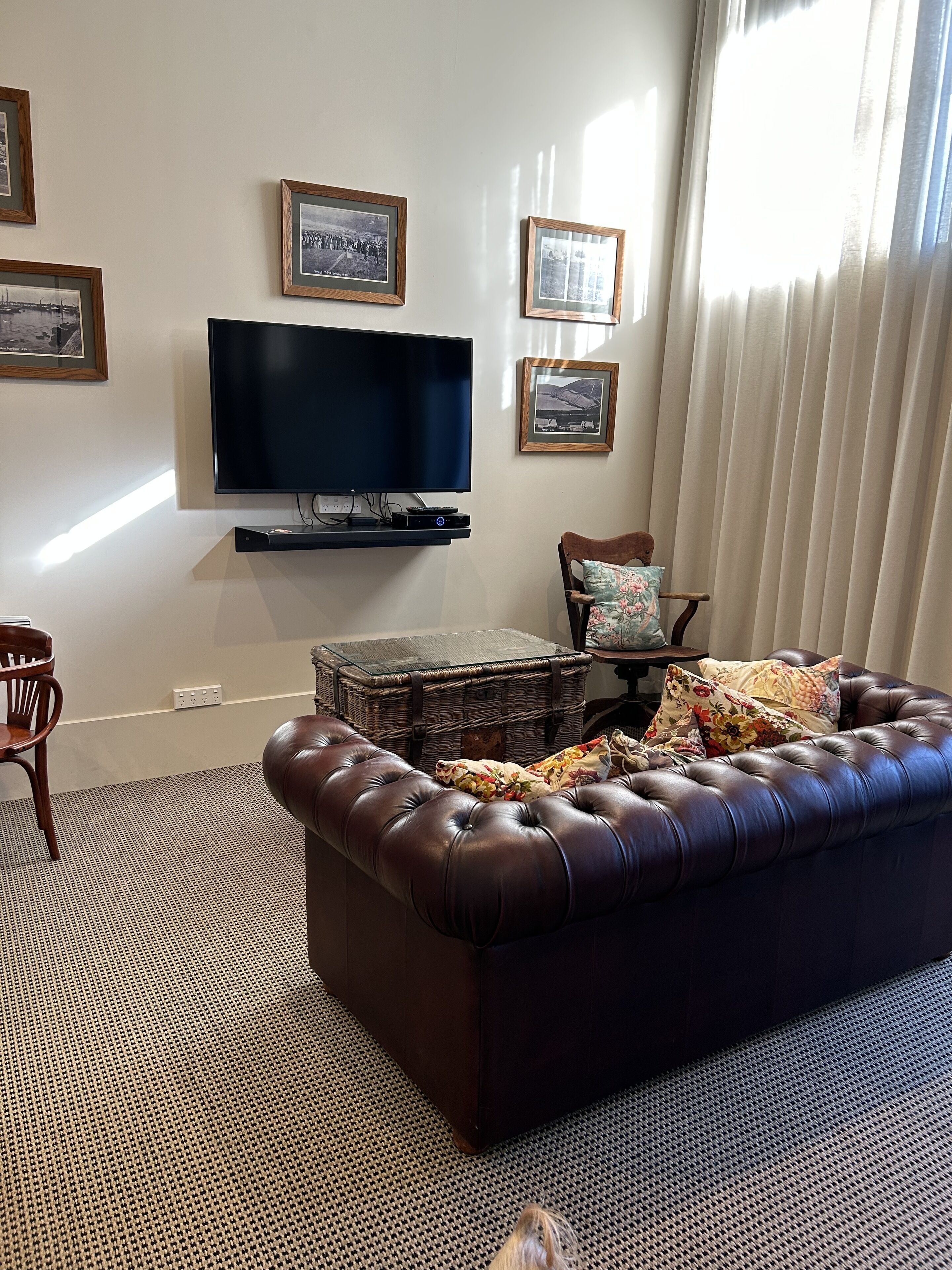 A living room with a large flat-screen TV, a brown leather sofa, a wicker side table, and a patterned rug.