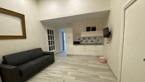 Una sala de estar moderna con un sofá gris, una cocina con armarios blancos y un televisor, y un pasillo que conduce a otra habitación.