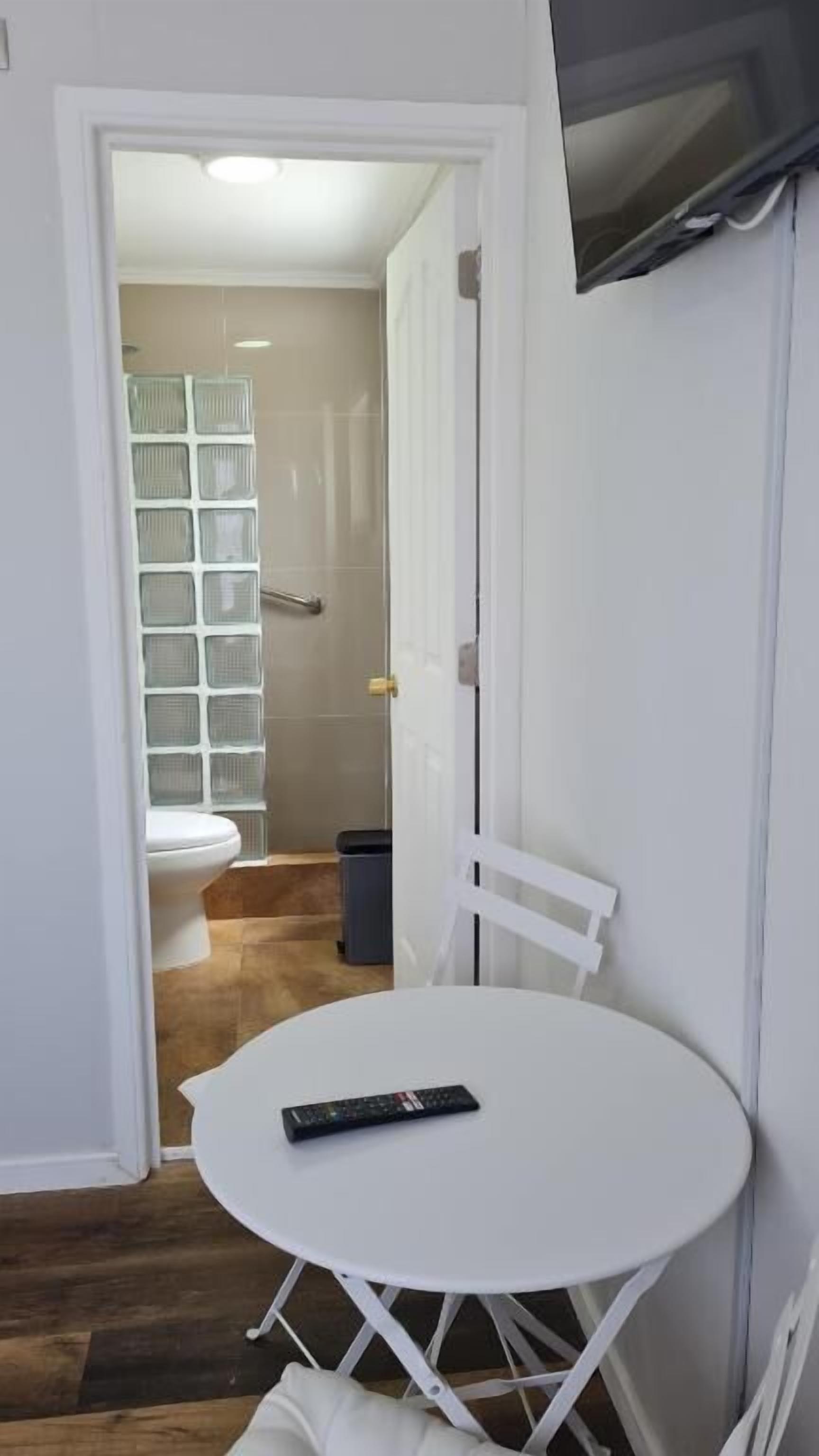 A small room with a white table and chairs, a remote control on the table, a white door, and a view into a bathroom with a toilet and a shower.