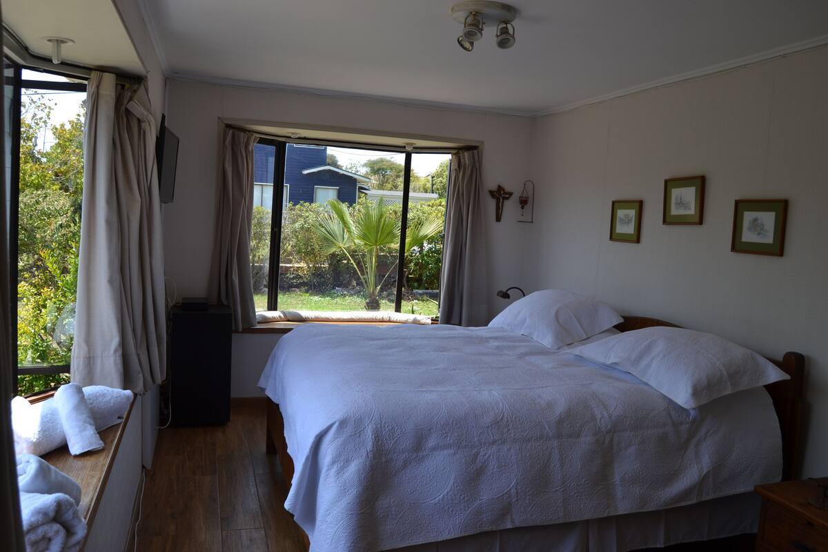 A bedroom with a large bed, a window with curtains, a wooden floor, and a view of greenery outside.