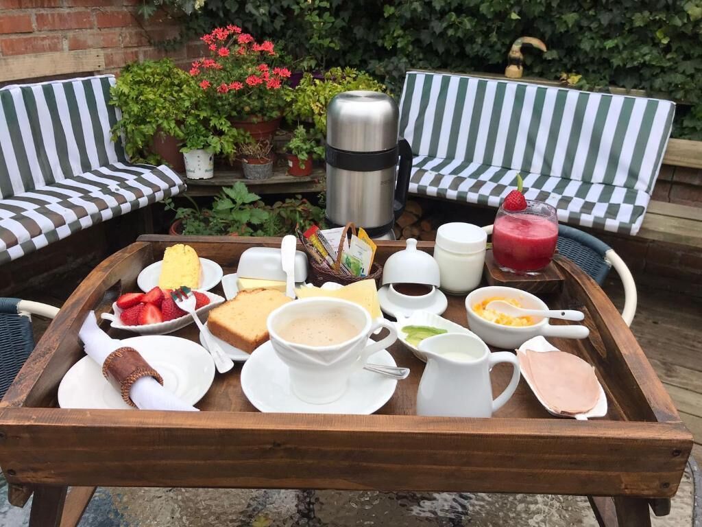 A tray with breakfast items including toast, fruit, and beverages, set on a wooden table outdoors with striped chairs and potted plants in the background.
